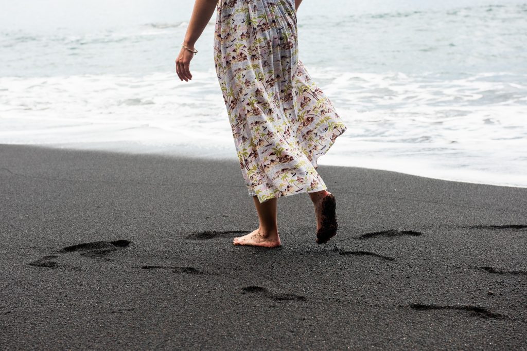 Woman from torso down walking barefoot on black sand beach wearing floral dress.
